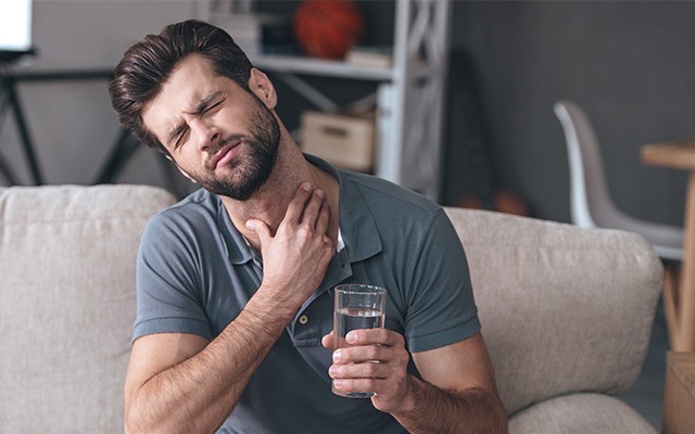 Ein Mann hält ein Glas Wasser und fasst sich mit schmerzverzerrtem Gesicht an den Hals.