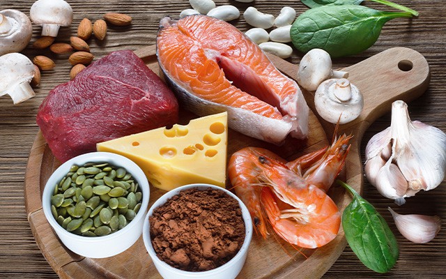 Verschiedene Lebensmittel, darunter Fleisch, Fisch, Käse und Gemüse, liegen auf einem Holzbrett.