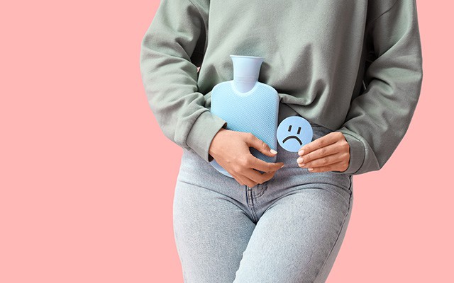 Person hält eine Wärmflasche auf den Unterbauch und ein trauriges Symbol in der Hand vor rosa Hintergrund.