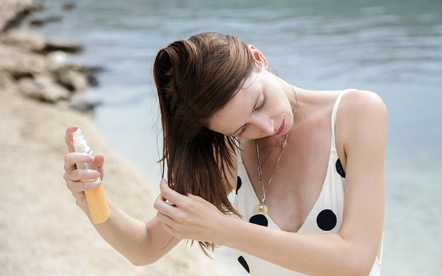 Eine Frau sprüht ein Produkt auf ihr Haar am Strand.