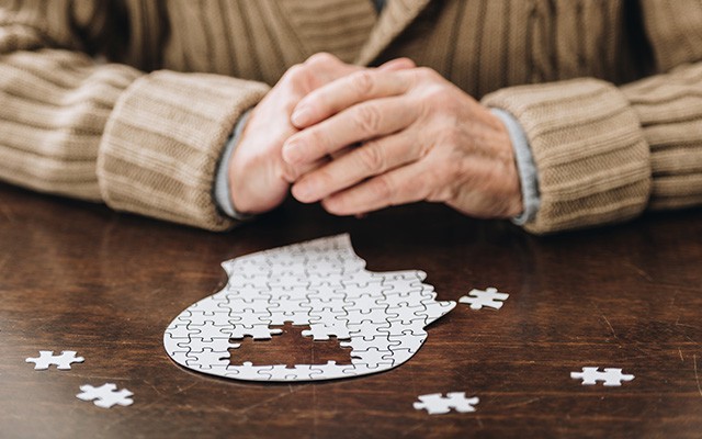 Ältere Person sitzt vor einem unvollständigen Puzzle in Kopf-Form auf einem Tisch.