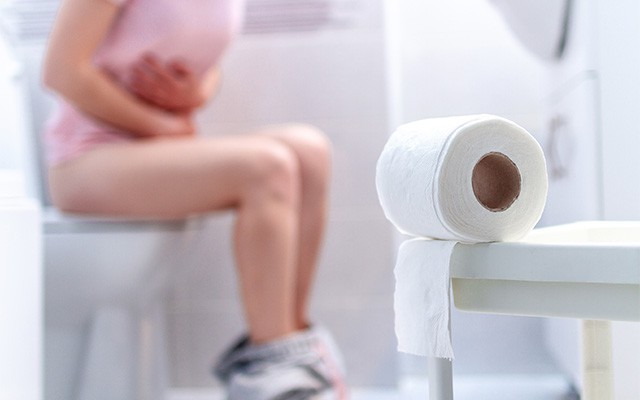 Person sitzt mit Bauchschmerzen auf der Toilette, daneben liegt Toilettenpapier.