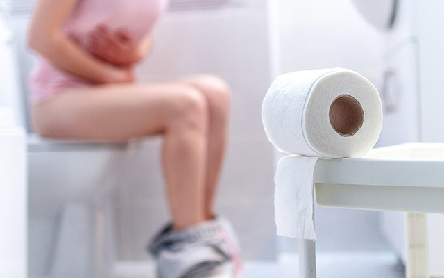 Person sitzt auf der Toilette und hält den Bauch, daneben eine Toilettenpapierrolle.