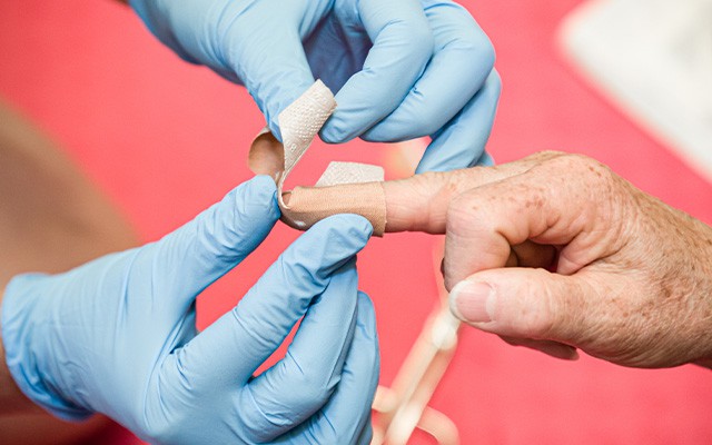 Eine Person mit blauen Handschuhen legt einem anderen eine Bandage um den Finger.