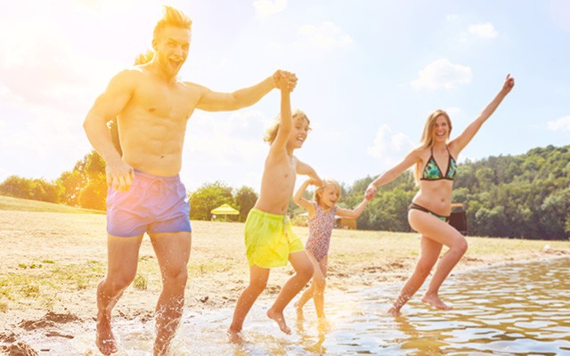 Familie läuft lachend durch flaches Wasser an einem sonnigen Badesee.