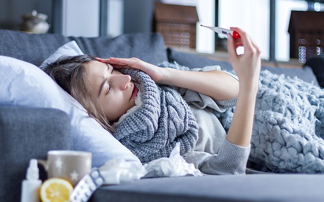 Frau liegt krank auf der Couch und hält ein Thermometer in der Hand.