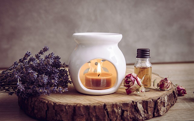 Weiße Duftlampe mit Teelicht, Fläschchen Öl und Lavendel auf Holzscheibe.