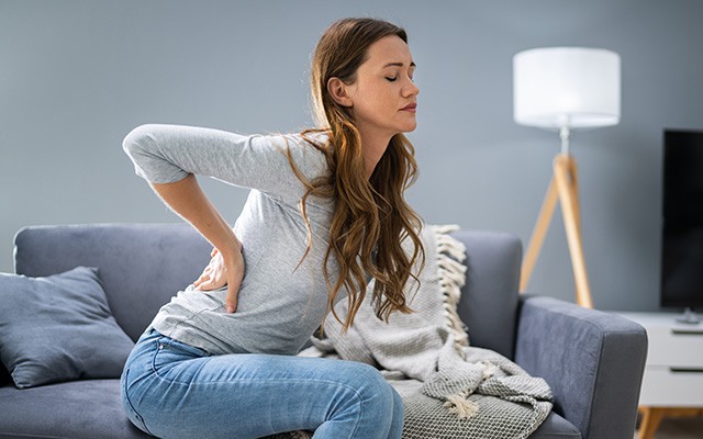 Eine Frau sitzt auf dem Sofa und hält ihren schmerzenden Rücken.