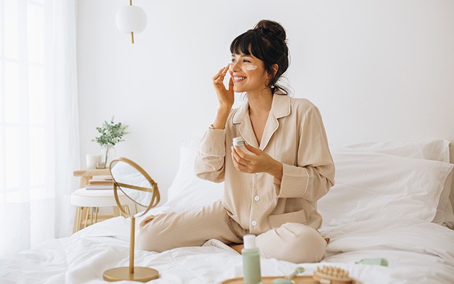 Eine Frau sitzt lächelnd in Pyjama auf dem Bett und cremt ihr Gesicht ein.