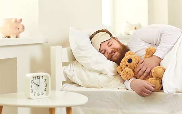 Ein Mann schläft lächelnd im Bett mit einer Schlafmaske und einem Teddybär.