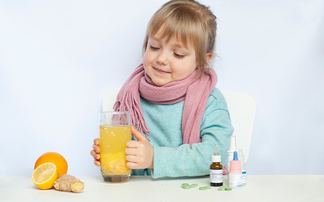 Ein Kind mit Schal hält ein Glas Saft, umgeben von Medikamenten, Zitrone und Ingwer.