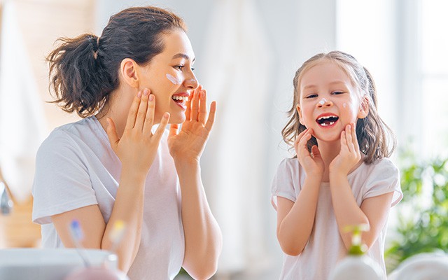 Mutter und Kind tragen gemeinsam Hautcreme auf und lachen im Badezimmer.