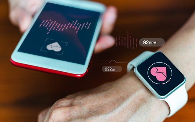 Eine Hand hält ein Smartphone, während am Handgelenk eine Smartwatch Herzfrequenz anzeigt.