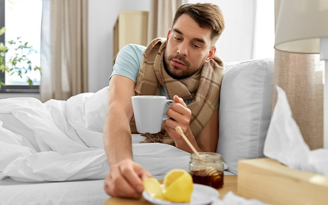 Ein Mann liegt krank im Bett und hält eine Tasse Tee mit Honig und Zitrone.