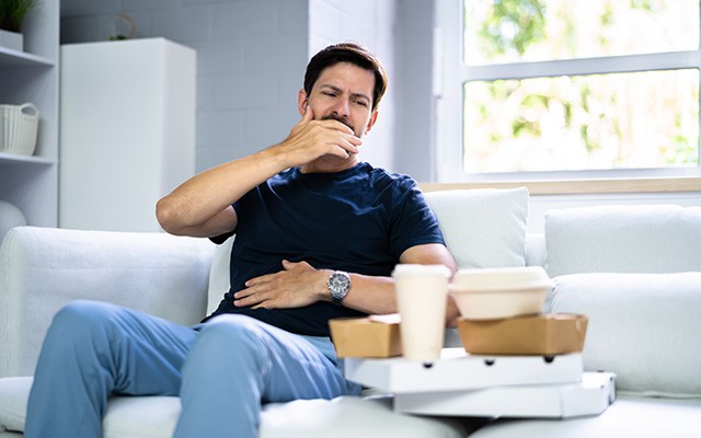 Ein Mann sitzt auf dem Sofa, hält seinen Bauch und gähnt.
