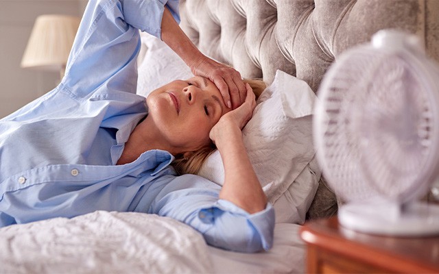 Eine Person liegt im Bett und fasst sich an die Stirn, ein Ventilator steht daneben.