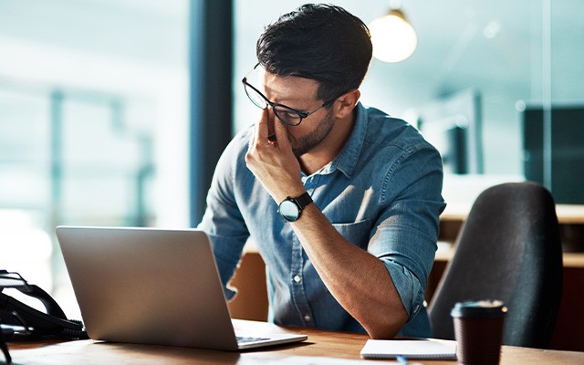 Ein Mann sitzt am Schreibtisch und reibt sich die Augen vor einem Laptop.