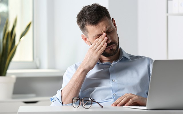 Ein Mann sitzt am Schreibtisch und reibt seine Augen müde.