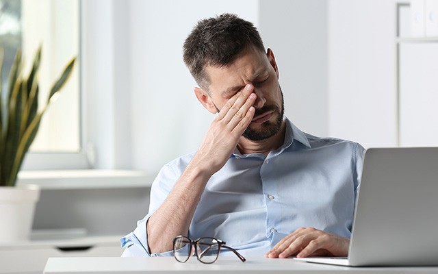Ein Mann sitzt am Tisch und reibt sich müde die Augen vor einem Laptop.