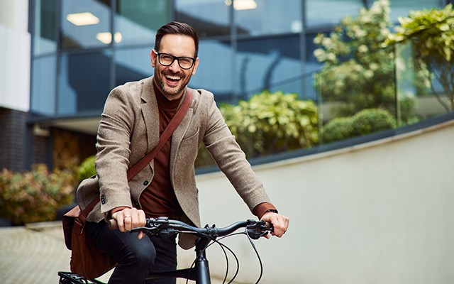 Ein Mann mit Brille lächelt und fährt Fahrrad vor einem Gebäude.