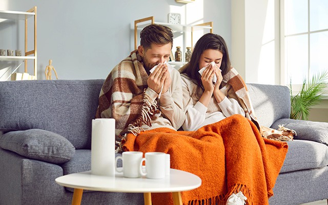 Ein Mann und eine Frau sitzen krank auf dem Sofa und schnäuzen sich die Nase.