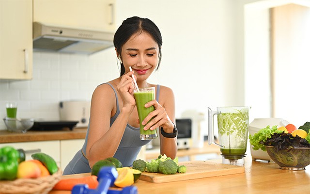 Eine Frau trinkt in der Küche einen grünen Smoothie aus einem Glas mit Strohhalm.