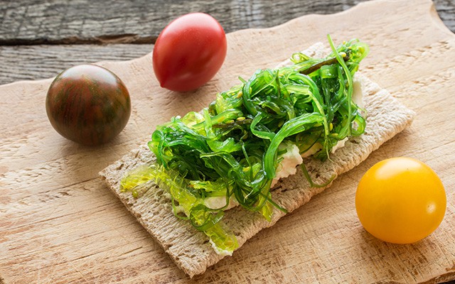 Knäckebrot mit Algensalat und drei bunten Tomaten auf Holzbrett.